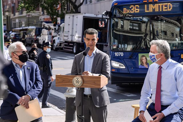 Ydanis Rodriguez, standing at a lectern and in front of a bus on a sidewalk, speaks with Mayor de Blasio on the right